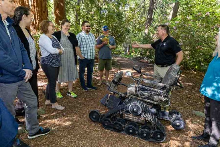 Representatives of the David's Chair nonprofit talk about the original chair created for use by disabled individuals on surfaces that are unusable by traditional wheelchairs during a grant presentation gathering in Lithia Park earlier this year. Ashland.news photo by Bob Palermini